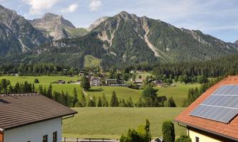 A picturesque landscape with green meadows and majestic mountains in the background. In the foreground, some houses are visible, including one with solar panels.