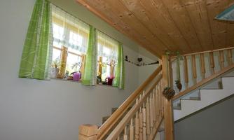 A bright hallway with a wooden staircase and large windows. The windows are decorated with green curtains and there are plants on the windowsill.