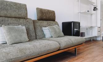 A modern living room with a comfortable gray sofa and decorative pillows. In the background, there is a black refrigerator and a shelf.