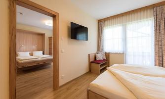 A cozy bedroom with wooden flooring and warm colors. In the background, there is a second room with two beds and a television visible.