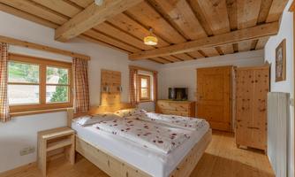 A cozy bedroom with wooden ceilings and furniture. Two beds with a floral bedspread and large windows let in plenty of daylight.