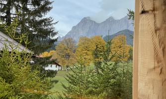 A tranquil landscape with colorful autumn trees and majestic mountains in the background. The scene is enveloped by green fir trees and appears peaceful.