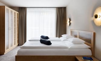A modern bedroom with a double bed and two large windows. The room is bright and furnished in a minimalist style.