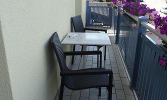 A balcony with two black chairs and a small table. The floor is tiled and in the background, plants are blooming.