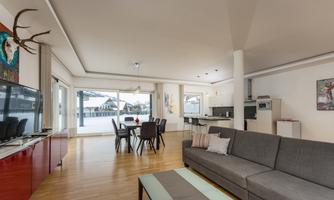 A modern living room with a large window front that offers a view of the surrounding mountains. The room is bright and minimally furnished with a gray sofa and a dining table.