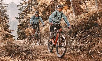 Two cyclists ride along a forest path through an autumn landscape. The trees are green and golden brown, and the surroundings appear peaceful and natural.