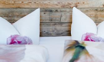 A cozy bed with white pillows and romantic, floral bedcovers. In the background, a wooden wall can be seen, creating a rustic atmosphere.