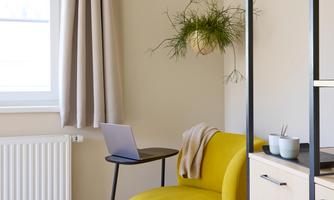 A cozy room with a yellow armchair and a small table. In the background, there are windows and neat shelves.