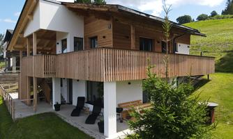 A modern house with wooden cladding and a spacious balcony. It is located in a green landscape with trees and meadows.