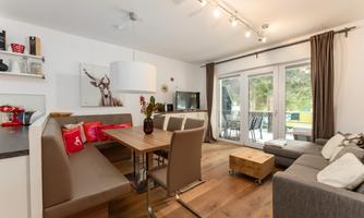 A modern apartment with a cozy dining area and an inviting living space. Large windows provide a view of the terrace and the green surroundings.