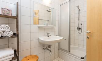 A modern bathroom with white tiles and a shower. There is a sink, towels, and a small wooden chair.