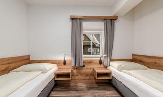 A modern bedroom with two single beds and wooden furniture. The room has a window with curtains and is bright and inviting.