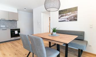 A modern dining room with a wooden table and gray benches. The kitchen in the background is bright and functionally designed.