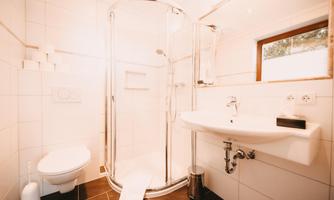 A modern bathroom with a shower cubicle, a wash basin, and a toilet. It is bright and clean with white tiles and a window that lets in daylight.