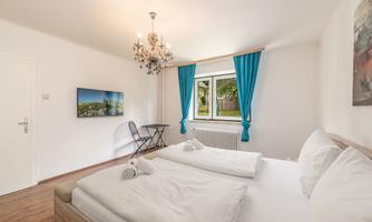 A bright bedroom with two beds, a cozy table, and a television. Large windows with blue curtains let in plenty of daylight.