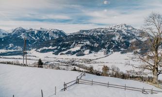 A winter mountain landscape with snow-covered hills and high mountains in the background. The sky is partly cloudy and conveys a calm atmosphere.