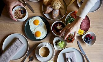 A lavish breakfast table with scrambled eggs, fruit, muesli, and various baked goods. Hands reach for the treats, creating an inviting atmosphere.