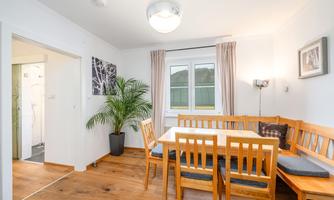 A bright dining room with a cozy seating area and a wooden table. Next to the table stands a large plant, and the window offers a view outside.