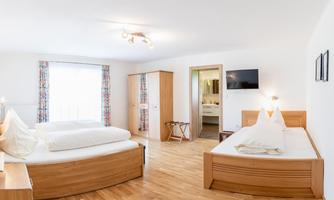 A bright and modern bedroom with two beds and wooden flooring. There is a wardrobe and a television, as well as large windows for plenty of daylight.