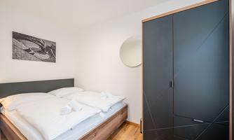 A modern bedroom with a double bed and white bedcovers. On the wall hangs a picture and a round mirror, next to the bed is a black wardrobe.
