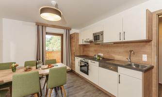 A modern kitchen with wood details and light furniture. The dining area is equipped with a table and green chairs.