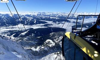 A cable car moves over snow-covered mountains with a clear blue sky in the background. From the height, there is a breathtaking view of the valley and the surrounding peaks.