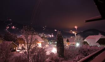 A snowy landscape at night with illuminated houses. In the background, light bursts and snow-covered hills are visible.