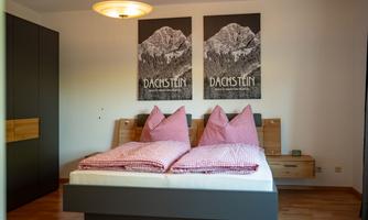 A cozy bedroom with a large bed and red checkered pillows. On the wall, two pictures of the Dachstein mountains are hanging.