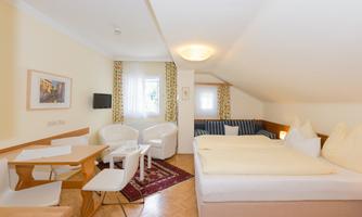 A cozy room with a double bed, a small table, and two chairs. Large windows provide plenty of daylight and a pleasant atmosphere.