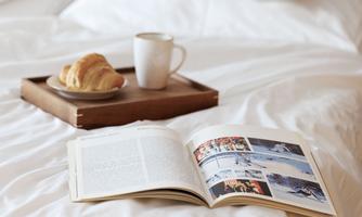 A cozy bed with an open book, a cup of coffee, and a croissant on a tray. The surroundings radiate relaxation and coziness.