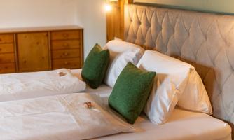 A cozy bedroom with two beds and green pillows. The walls are light, and a wooden wardrobe is visible in the background.