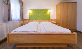 A simple bedroom with a double bed and a wooden wardrobe. The walls are light and decorated with a green accent wall.