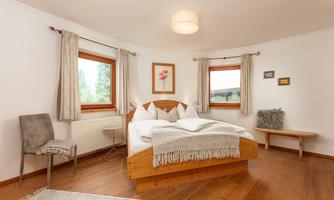 A cozy bedroom with a large bed and hardwood floor. The walls are painted light, and there are windows with curtains that provide natural light.