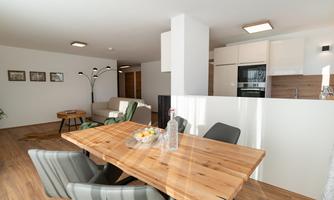 A modern, bright apartment with a wooden dining table and cozy chairs. In the background, a kitchen and a living room with a sofa can be seen.