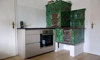 A modern kitchen with a shiny silver stove and a traditional green tiled stove. The room features bright wooden floors and a minimalist design.