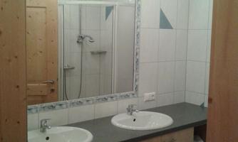 A modern bathroom with two sinks and a mirror. The walls are tiled in white and there is a shower in the background.