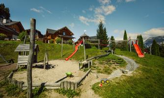 A playground with a sand area, slides, and a small swing. In the background, there are houses and trees.