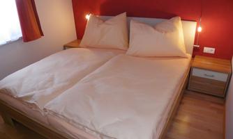 A cozy bedroom with a double bed and soft pillows. The wall is painted red and a nightstand is next to it.