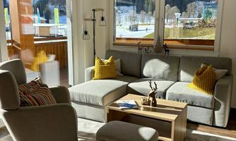 A cozy living room with a bright seating area and a wooden table. Large windows offer a view of the surrounding nature.