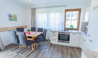 A modern kitchen with a wooden dining area and black chairs. Large windows provide plenty of daylight in the room.