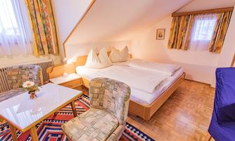 A cozy bedroom with two beds and bright pillows. There is a table and chairs as well as colorful curtains and a carpet.