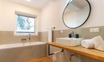 A modern bathroom with a bathtub and a sink on a wooden shelf. Large windows provide plenty of natural light and a welcoming atmosphere.