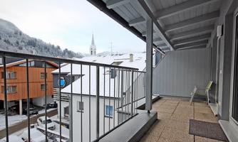 A balcony with a view of snow-covered roofs and surrounding mountains. In the background, a church tower is visible.