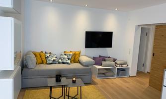 A modern living room with a bright couch and colorful cushions. A television hangs on the wall, and the floor is covered with a yellow rug.