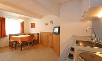 A cozy kitchenette with a dining area and a small television. The room is bright and inviting, with windows and orange curtains.