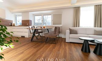 A modern living room with a bright, open space. It includes a dining area, a couch, and large windows that let in plenty of daylight.