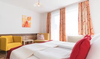 A bright room with a large bed and red pillows. In the background, there is a cozy seating area and a large window front with curtains.