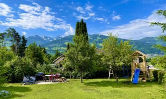 A green garden landscape with play equipment and trees. In the background, the mountains can be seen under a blue sky.