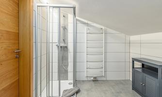 A modern bathroom with a shower and a towel. The walls are tiled with white tiles, and a gray cabinet stands in the corner.