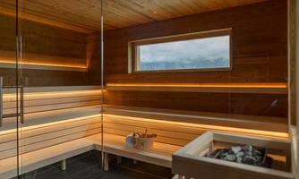 A modern sauna room with wooden lounging areas and soft lighting. A large window provides a view of a mountainous landscape.
